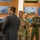 Department of War Delegation visits Poland A civilian in a business suit is briefing or presenting to a group of U.S. Air Force military personnel in a room decorated with aircraft photographs and aviation imagery on brick walls. The setting appears to be a military facility, likely a briefing room or heritage room, with multiple uniformed service members listening attentively. Personnel are wearing a mix of OCP (Operational Camouflage Pattern) uniforms and a green flight suit.