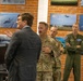 A civilian in a business suit is briefing or presenting to a group of U.S. Air Force military personnel in a room decorated with aircraft photographs and aviation imagery on brick walls. The setting appears to be a military facility, likely a briefing room or heritage room, with multiple uniformed service members listening attentively. Personnel are wearing a mix of OCP (Operational Camouflage Pattern) uniforms and a green flight suit.