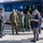 Department of War Delegation visits Poland A group of military personnel in various uniforms and a civilian in a suit are gathered outside near a large IVECO Crossway bus on a paved area. The scene appears to be a formal or official meeting, possibly a briefing or reception at a military or government facility. Personnel are wearing different camouflage and flight suit uniforms, suggesting a multinational or inter-service gathering.
