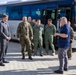 A group of military personnel in various uniforms and a civilian in a suit are gathered outside near a large IVECO Crossway bus on a paved area. The scene appears to be a formal or official meeting, possibly a briefing or reception at a military or government facility. Personnel are wearing different camouflage and flight suit uniforms, suggesting a multinational or inter-service gathering.
