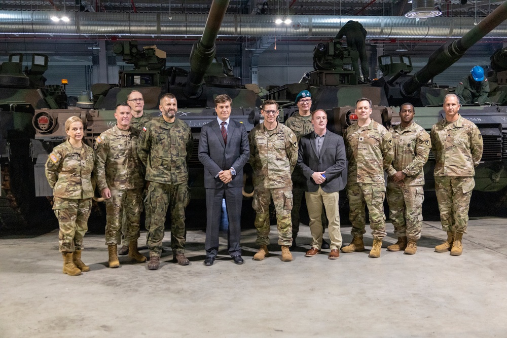 A group of approximately twelve individuals, including military personnel in camouflage uniforms and two civilians in business attire, pose for a formal group photograph inside a military vehicle storage facility. Two large main battle tanks, appearing to be M1 Abrams, are visible in the background. The setting appears to be an indoor hangar or depot with industrial lighting and ventilation systems overhead.