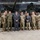 Department of War Delegation visits Poland A group of approximately twelve individuals, including military personnel in camouflage uniforms and two civilians in business attire, pose for a formal group photograph inside a military vehicle storage facility. Two large main battle tanks, appearing to be M1 Abrams, are visible in the background. The setting appears to be an indoor hangar or depot with industrial lighting and ventilation systems overhead.