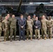 A group of approximately twelve individuals, including military personnel in camouflage uniforms and two civilians in business attire, pose for a formal group photograph inside a military vehicle storage facility. Two large main battle tanks, appearing to be M1 Abrams, are visible in the background. The setting appears to be an indoor hangar or depot with industrial lighting and ventilation systems overhead.
