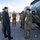 Department of War Delegation visits Poland A civilian in a dark business suit is speaking with a group of military personnel in MultiCam/OCP camouflage uniforms on what appears to be a military airfield or tarmac. A large olive-drab military tactical vehicle is visible on the right side of the frame. The setting suggests an official visit, inspection, or briefing by a civilian official or dignitary.