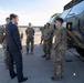 A civilian in a dark business suit is speaking with a group of military personnel in MultiCam/OCP camouflage uniforms on what appears to be a military airfield or tarmac. A large olive-drab military tactical vehicle is visible on the right side of the frame. The setting suggests an official visit, inspection, or briefing by a civilian official or dignitary.