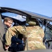 Two individuals are examining the cockpit area of a military helicopter, likely an Apache or similar attack helicopter. One person is wearing military camouflage (OCP/MultiCam pattern) uniform, while the other is dressed in civilian business attire. The interaction appears to be a briefing or demonstration, possibly during an official visit or inspection.