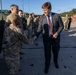A civilian in a dark suit and red striped tie is shaking hands with a soldier in military camouflage uniform at what appears to be a military installation or staging area. Multiple soldiers in camouflage uniforms are visible in the background alongside military vehicles. The setting appears to be an outdoor paved area with trees and storage containers visible in the background.