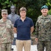 Three men pose for a photo in front of a bronze bust monument in an outdoor setting. Two of the men are in military uniforms — one wearing a U.S. Army uniform with the name tape 'MOUNTFORD' and a brigadier general star insignia, and the other wearing a Polish Army uniform with a green beret and Polish flag patch. The civilian in the center is dressed in a navy polo shirt and khaki trousers.