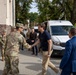 The image shows a formal greeting between a U.S. military officer in MultiCam/OCP camouflage uniform and a civilian man in casual attire on a paved outdoor area near a brick building. Several other military personnel in U.S. Army uniforms and what appears to be Polish military uniforms are present in the background. A white van and civilian vehicle are also visible, suggesting an official visit or delegation meeting.