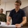 Department of War Delegation visits Poland A civilian man wearing glasses and a navy polo shirt is signing or writing in a large document binder at a desk. In the background, a military personnel wearing Polish Army camouflage uniform with a visible Polish flag patch stands and observes. The setting appears to be an official office or administrative room.
