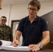 A civilian man wearing glasses and a navy polo shirt is signing or writing in a large document binder at a desk. In the background, a military personnel wearing Polish Army camouflage uniform with a visible Polish flag patch stands and observes. The setting appears to be an official office or administrative room.