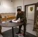 Three military personnel are gathered around a round table in what appears to be a military office or reception area. A soldier in a distinctive camouflage pattern — consistent with Polish Armed Forces uniform — is signing a guest book or official document, while two U.S. Army soldiers in MultiCam/OCP uniforms observe. The setting includes military insignia, unit patches, and a poster on the wall suggesting a U.S. Army unit headquarters.