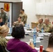 A military briefing or meeting is taking place in a conference room, with multiple uniformed personnel seated around a dark oval table. The group includes both military officers in camouflage uniforms and at least two civilians. Name placards are visible on the table, and one officer appears to be speaking or presenting to the group.