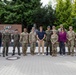 A group of approximately 16 individuals, mostly in military uniforms, pose for a formal group photograph in front of a sign reading 'Camp Kościuszko'. The group includes personnel in US Army combat uniforms (OCP pattern), at least one Polish military uniform, one individual in Air Force flight suit, and two civilians in casual attire. The setting appears to be a military installation entrance or memorial area.