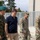 Department of War Delegation visits Poland The image shows a group of four individuals walking together in what appears to be a military installation or base. The group includes one Polish military officer in camouflage uniform with a green beret, one civilian in a navy polo shirt, and two U.S. Army soldiers in MultiCam (OCP) uniforms. The setting suggests an official visit or coordination meeting between allied military and civilian personnel.