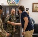 The image shows a meeting or greeting between military personnel and a civilian in what appears to be a military office or reception area. A Polish soldier and a US Army soldier are present, with the Polish soldier shaking hands with a civilian man in casual attire. The setting includes military insignia, framed artwork, and display cases on the walls.