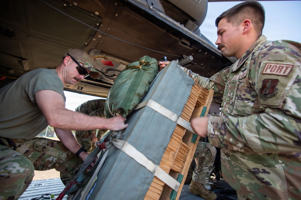 Two U.S. Air Force personnel in OCP (Operational Camouflage Pattern) uniforms are loading or unloading cargo onto/from a military aircraft. The soldier on the right wears a 'PORT' tab patch, indicating a Port Dawg or aerial port specialist role. They are handling a strapped cargo bundle on what appears to be a standard military cargo pallet.