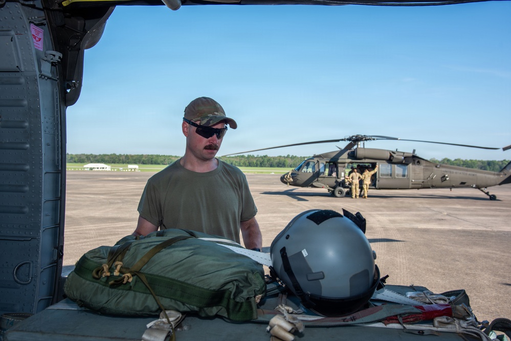A military service member in olive drab t-shirt and multicam cap is loading or organizing equipment inside what appears to be a military helicopter. In the foreground, a flight helmet and green equipment bags are visible on a surface. In the background, a UH-60 Black Hawk helicopter is parked on a tarmac with personnel working around it.
