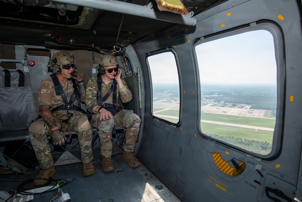 Two military personnel in full combat gear are seated inside a military helicopter in flight, looking out a window at an airfield below. Both individuals are wearing multicam uniforms, tactical helmets, sunglasses, and communication headsets. The aerial view through the window reveals a military airfield with runways, taxiways, and parked aircraft visible on the ground.