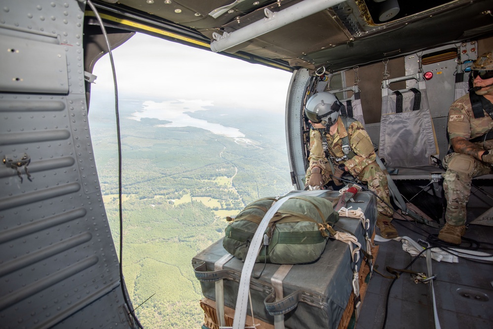 Military personnel are positioned inside an open-doored aircraft at altitude, preparing to deploy or push out cargo/equipment. The aircraft interior shows riveted metal construction typical of military transport helicopters or aircraft, with the open door revealing a sweeping aerial view of forested terrain and a river or lake below. At least two personnel in military camouflage (OCP/Multicam) and flight gear are visible managing the operation.