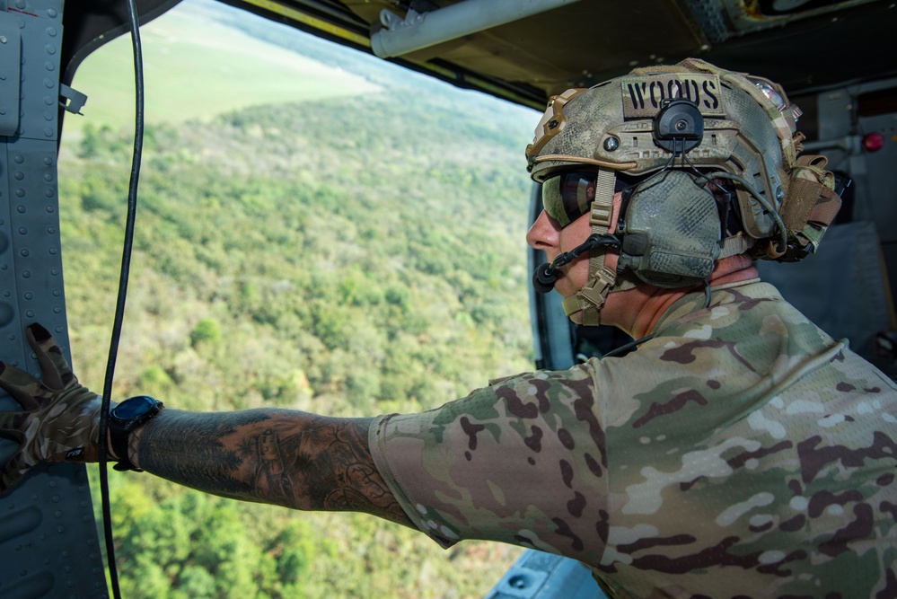 A military personnel member identified as 'WOODS' on their helmet is looking out from an open door of a military helicopter over a densely forested landscape. The individual is wearing full tactical gear including a multicam helmet with communications equipment, multicam uniform, and tactical gloves. The aerial perspective suggests the aircraft is flying at a moderate altitude over a green, tree-covered terrain.