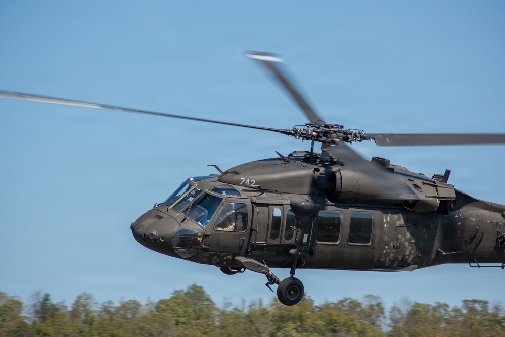 A military utility helicopter, identifiable as a UH-60 Black Hawk, marked with the number '742' is captured in low-altitude flight against a clear blue sky. The helicopter is painted in a flat olive drab/dark green military finish and appears to be in active flight with rotor blades in motion. Treeline vegetation is visible in the background below the aircraft.