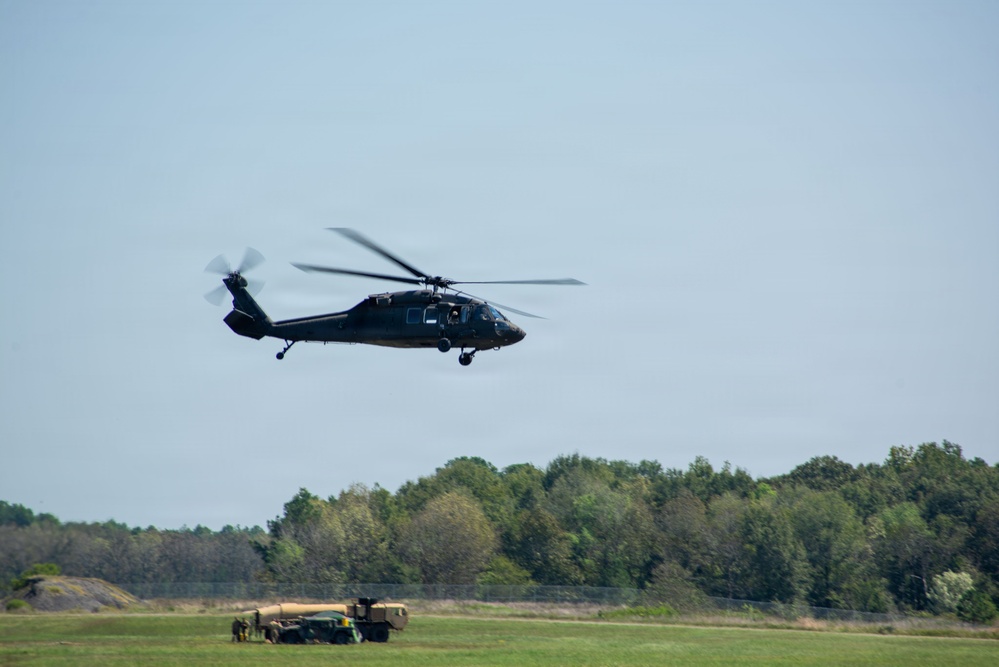 A dark military helicopter, consistent with a UH-60 Black Hawk, is captured in low-altitude flight over an open grassy field. In the background, military ground vehicles and personnel are visible near a treeline. The image appears to depict a military training or operational exercise.