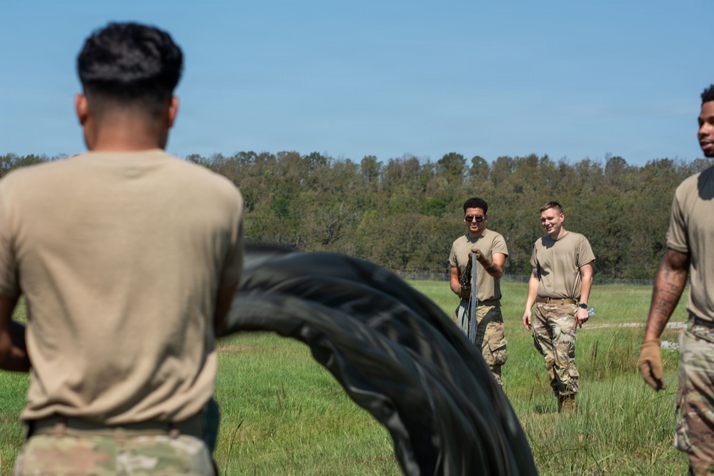 A group of military personnel in tan/camouflage uniforms are conducting outdoor physical training on a grassy field. The activity appears to involve battle rope or heavy rope exercises, with one individual in the foreground flipping or waving a large black rope/tire. Two soldiers in the background are smiling and carrying additional equipment.