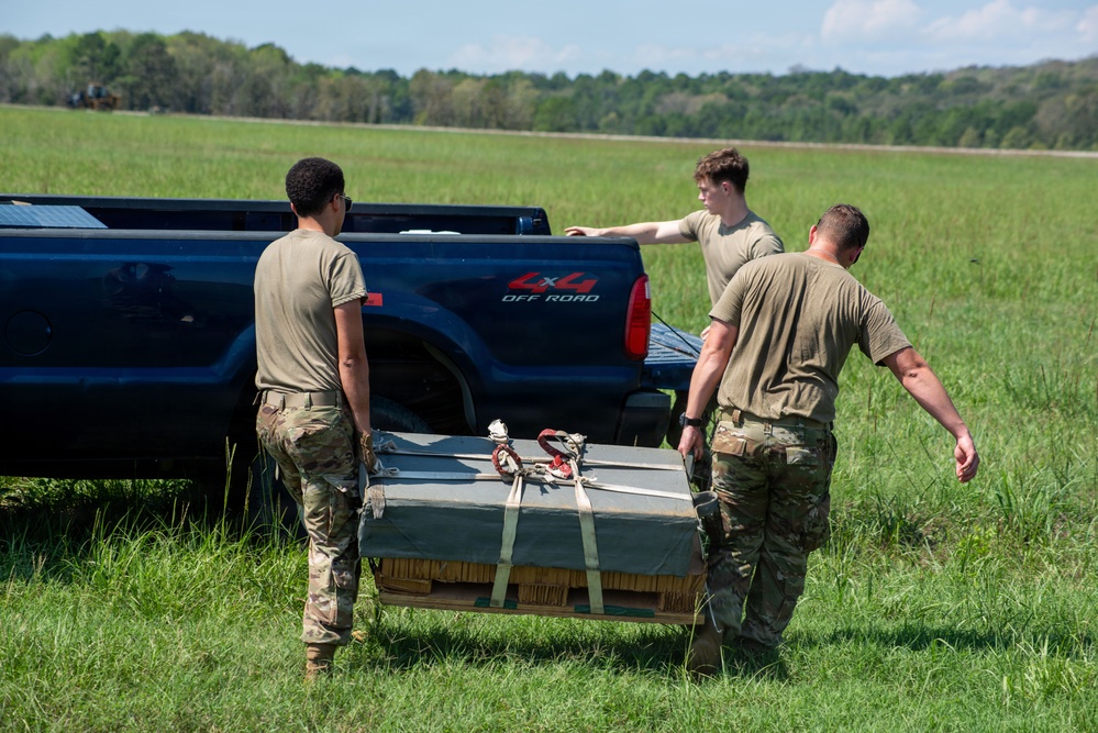Three military personnel in OCP (Operational Camouflage Pattern) uniforms are loading or unloading a strapped pallet or flat cargo item onto/from the bed of a dark blue 4x4 off-road pickup truck in an open grassy field. The individuals appear to be conducting a field logistics or training exercise. The setting is a rural, open area with tree lines visible in the background.