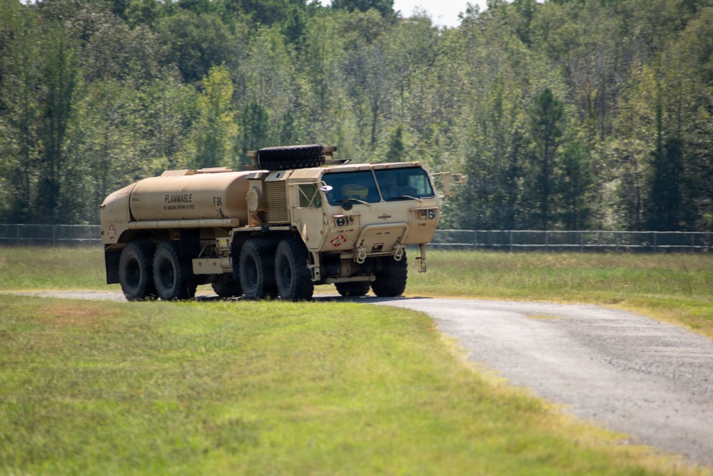 A desert tan military fuel tanker truck, likely an HEMTT (Heavy Expanded Mobility Tactical Truck) with a fuel pod, is traveling along a gravel road bordered by green grass and dense tree line. The tank is marked with 'FLAMMABLE' and hazmat placards, indicating it carries fuel or other flammable liquid. The setting appears to be a military training installation or base.
