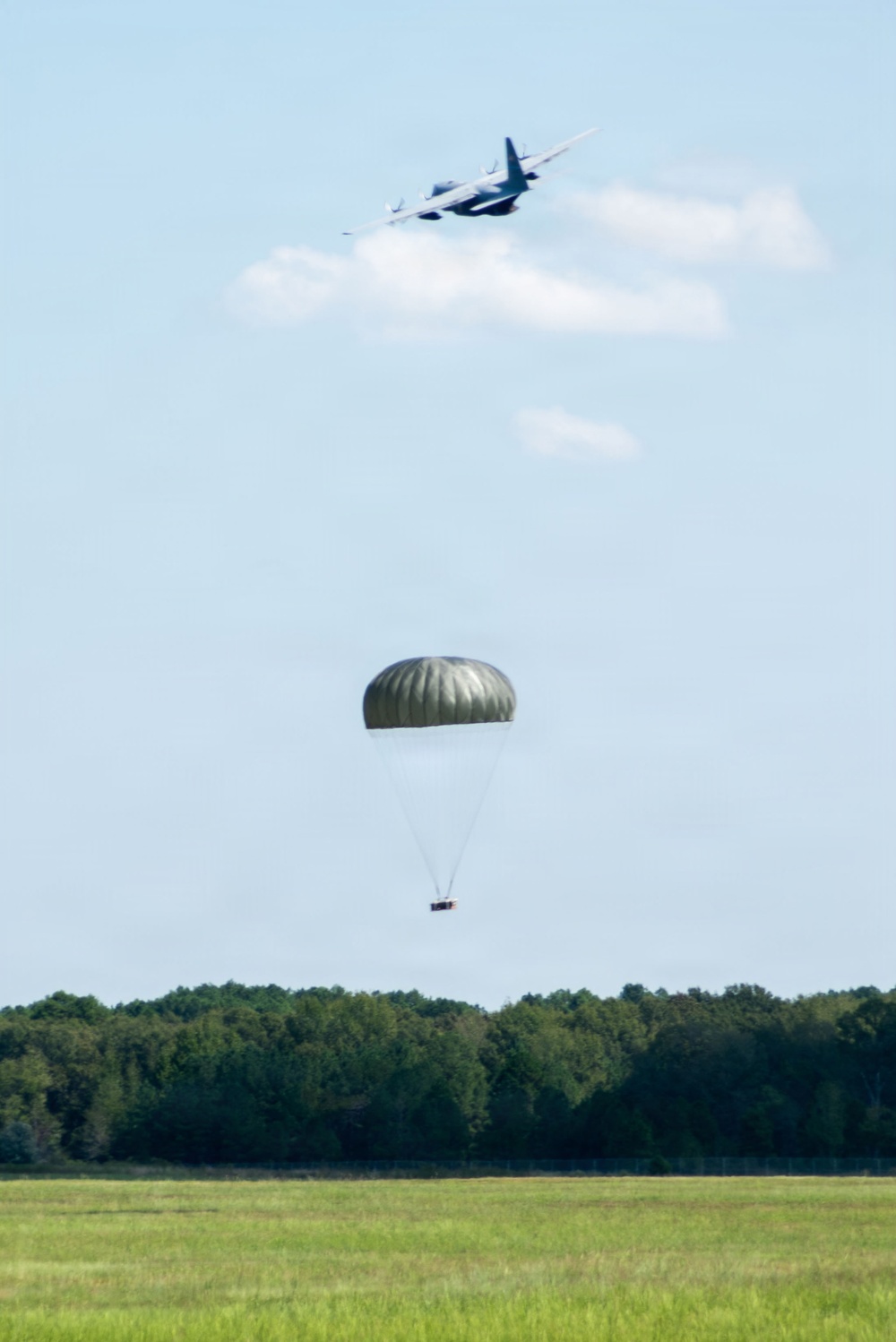 A military cargo aircraft, consistent with a C-130 Hercules, is flying overhead after releasing a supply bundle suspended beneath a large olive-drab parachute. The parachute and its attached cargo are descending toward an open grassy field bordered by a tree line. This appears to be a military airdrop or aerial delivery exercise.
