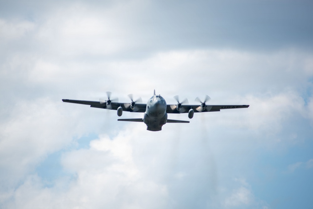 A four-engine turboprop military transport aircraft is photographed head-on in flight against a partly cloudy sky. The aircraft displays a gray military paint scheme and all four propellers are visibly spinning. The low-angle shot captures the aircraft approaching the camera with its wings fully extended.
