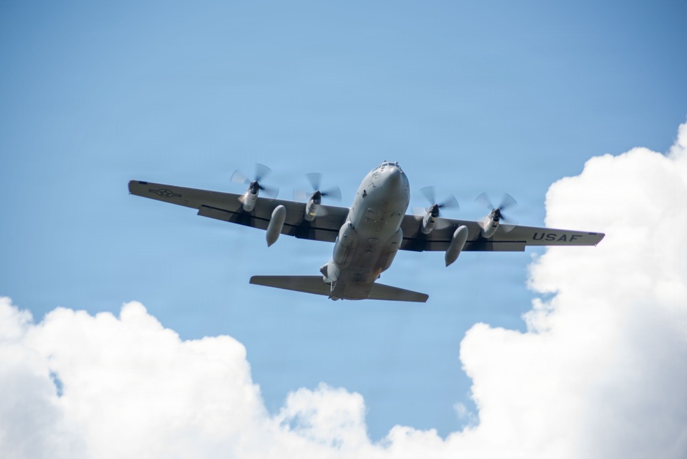 A United States Air Force (USAF) military transport aircraft, identifiable as a C-130 Hercules, is photographed from below as it flies overhead. The four-engine turboprop aircraft is captured in flight against a partly cloudy blue sky. The 'USAF' marking is clearly visible on the fuselage.