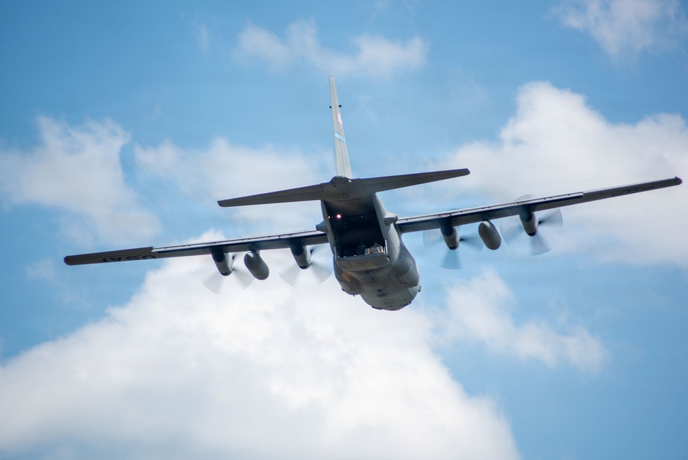 A large military turboprop transport aircraft is photographed from below and behind as it flies overhead against a partly cloudy blue sky. The aircraft's rear cargo ramp appears to be open or partially open, revealing the interior. The image is taken from a low angle, providing a clear view of the underside, wings, and tail assembly.