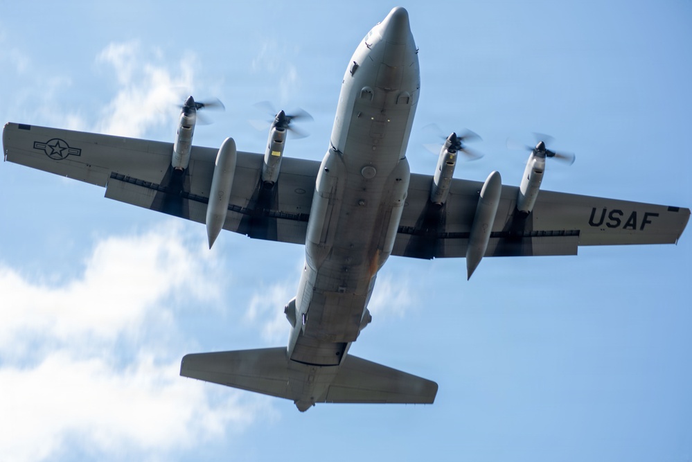 A United States Air Force (USAF) military transport aircraft is photographed from directly below as it flies overhead. The aircraft is a four-engine turboprop, consistent with a C-130 Hercules variant, displaying USAF markings and a roundel on the wing. The image is taken from a ground-level upward perspective against a partly cloudy blue sky.