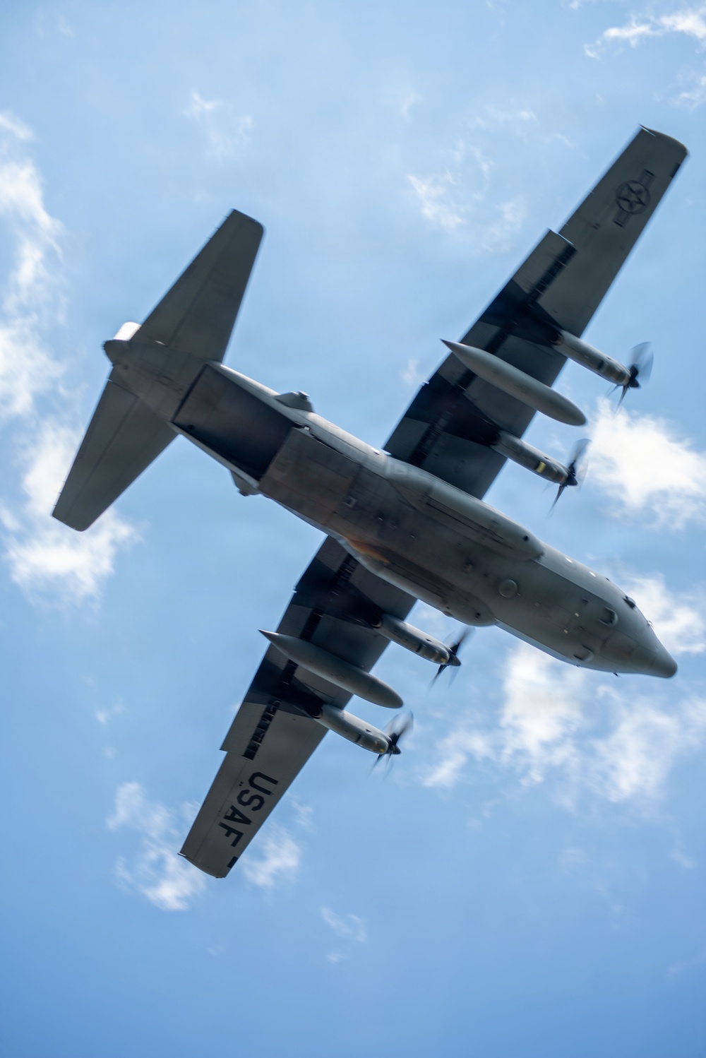 A USAF military transport aircraft, identifiable as a C-130 Hercules variant, is photographed from below at a banking angle against a partly cloudy blue sky. The underside of the aircraft is fully visible, showing four turboprop engines with spinning propellers, wing-mounted external fuel tanks or pods, and standard military gray paint scheme. The 'USAF' marking is clearly visible on the lower wing surface along with a military roundel on the tail.