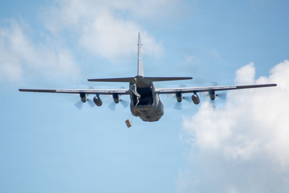 A military transport aircraft, consistent with a C-130 Hercules, is photographed from the rear during an airdrop operation. The aircraft's rear cargo ramp is open and a package or cargo bundle is visible being deployed beneath the fuselage. A figure, likely a loadmaster or jumpmaster, is visible standing at the open ramp.