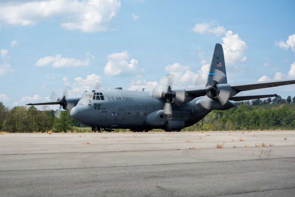 A U.S. Air Force C-130 Hercules military transport aircraft is taxiing or stationary on a tarmac with its four turboprop engines running, as indicated by the spinning propellers. The aircraft is painted in standard military gray livery with 'U.S. AIR FORCE' markings on the fuselage and an American flag on the tail. The setting appears to be a somewhat aged or lightly used airstrip surrounded by trees under a partly cloudy sky.