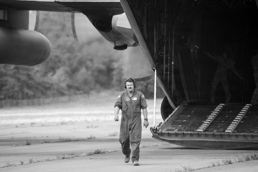 A black and white photograph showing a military aircrew member in a flight suit walking away from the open rear cargo ramp of a large military transport aircraft, likely a C-130 Hercules. The scene appears to be on an active airstrip or tarmac, with dust or exhaust visible in the background. A second figure is partially visible near the open cargo ramp.