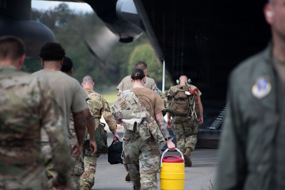 A group of military personnel in camouflage uniforms are boarding or loading equipment onto a large military transport aircraft, likely a C-130 Hercules, via its rear cargo ramp. The personnel are carrying bags, backpacks, and other equipment including a distinctive yellow and red container. A figure in a flight suit is visible in the foreground right.