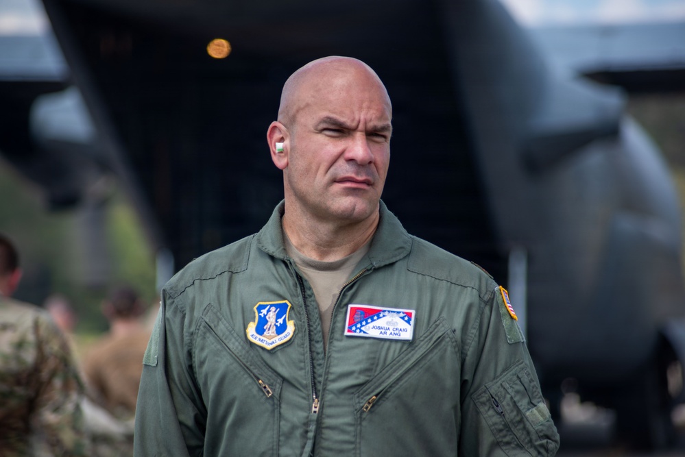 A bald male military personnel wearing an olive green flight suit stands in front of what appears to be a military aircraft. His name tag reads 'Joshua Craig' and his unit patch indicates 'Air National Guard' (AR ANG - Arkansas Air National Guard). He has a white earpiece in his right ear and appears to be observing something off-camera.