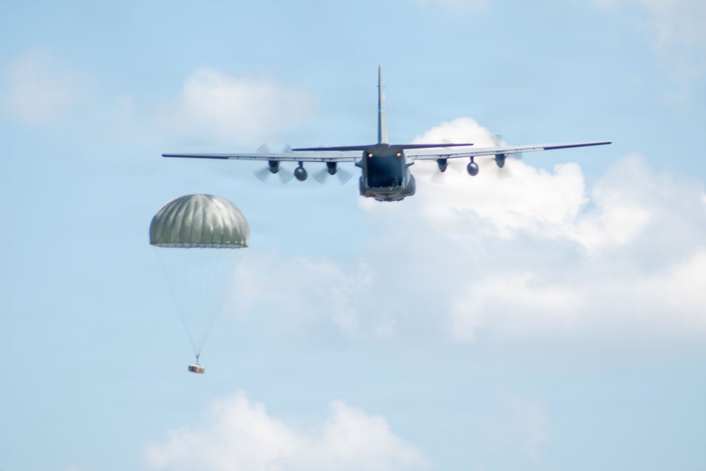 A military transport aircraft, consistent with a C-130 Hercules configuration, is photographed from the rear as it conducts an airdrop operation. A single olive-drab/green parachute has deployed to the left of the aircraft, suspending what appears to be a cargo bundle or supply package. The image was taken in daylight against a partly cloudy blue sky.
