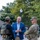 Three individuals are engaged in a conversation outdoors in what appears to be a public plaza or urban area. The group consists of a civilian in business casual attire flanked by a U.S. Marshal in tactical gear and a National Guard soldier in full military uniform. The setting suggests an official coordination or briefing meeting between law enforcement and military personnel.