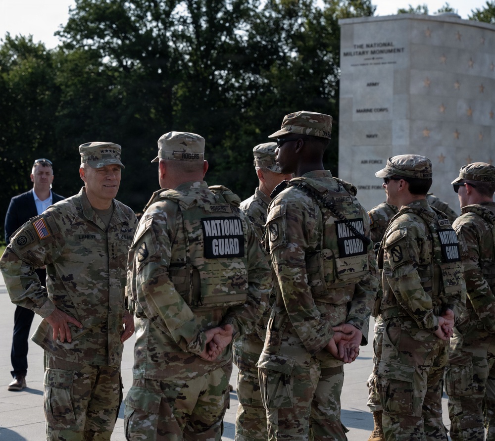 A group of National Guard soldiers in OCP (Operational Camouflage Pattern) uniforms are gathered at what appears to be the National Military Monument. A senior officer, identifiable by rank insignia on his cap, appears to be addressing or speaking with the assembled soldiers. The setting is an outdoor plaza with trees and a large stone monument visible in the background.