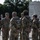 A group of National Guard soldiers in OCP (Operational Camouflage Pattern) uniforms are gathered at what appears to be the National Military Monument. A senior officer, identifiable by rank insignia on his cap, appears to be addressing or speaking with the assembled soldiers. The setting is an outdoor plaza with trees and a large stone monument visible in the background.