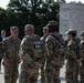 A group of National Guard soldiers in OCP (Operational Camouflage Pattern) uniforms are gathered at what appears to be the National Military Monument. A senior officer, identifiable by rank insignia on his cap, appears to be addressing or speaking with the assembled soldiers. The setting is an outdoor plaza with trees and a large stone monument visible in the background.