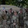 A group of four U.S. Army soldiers in MultiCam (OCP) camouflage uniforms pose for a photo in front of a tan military HMMWV (Humvee). A fifth soldier, with the name tape 'RAINES' visible, photographs them using a smartphone. One of the posing soldiers has the name tape 'WEIMER' visible, and the setting appears to be an urban environment.
