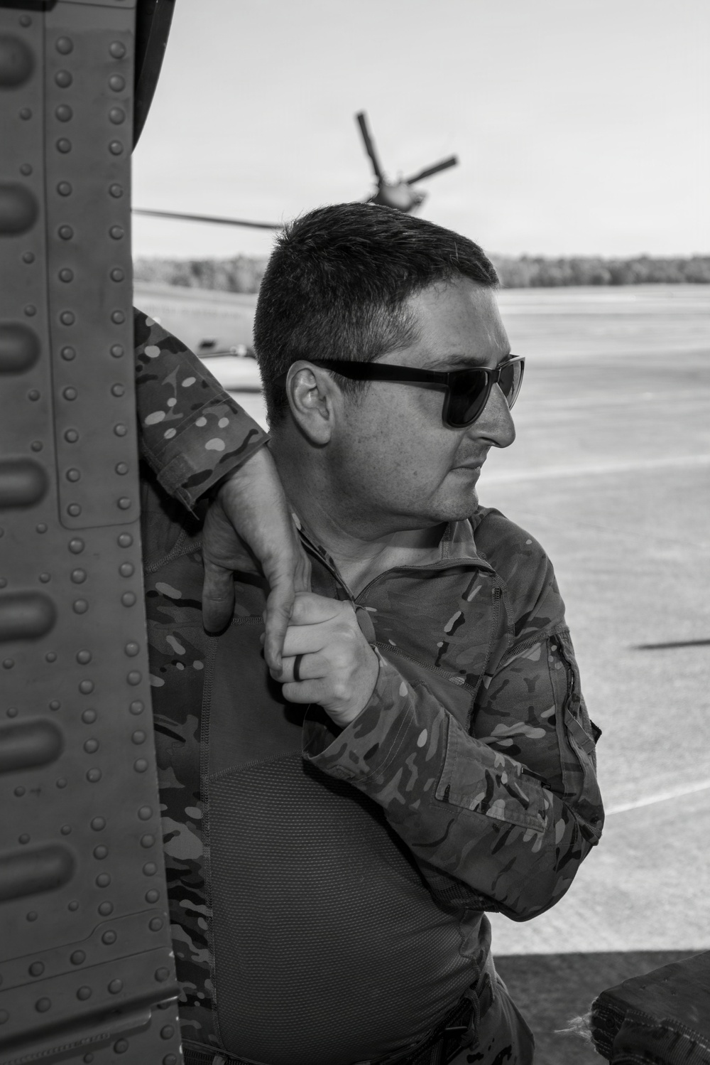 A black-and-white photograph of a male individual wearing camouflage military-style clothing and sunglasses, leaning against what appears to be a military aircraft or helicopter fuselage. He is looking to the right, and a helicopter with visible rotor blades is visible in the background on what appears to be a tarmac or airfield. The image has a professional, documentary-style composition.