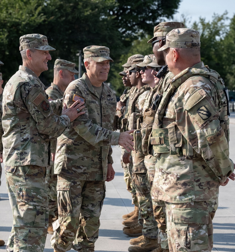 A senior U.S. Army officer wearing four-star general rank insignia is shaking hands with soldiers in a formal greeting or inspection ceremony. The general, whose name tape reads 'LANEYE' or similar, is accompanied by another high-ranking officer (visible two-star rank) as they move down a line of uniformed soldiers. The setting appears to be an outdoor military installation during daylight hours.