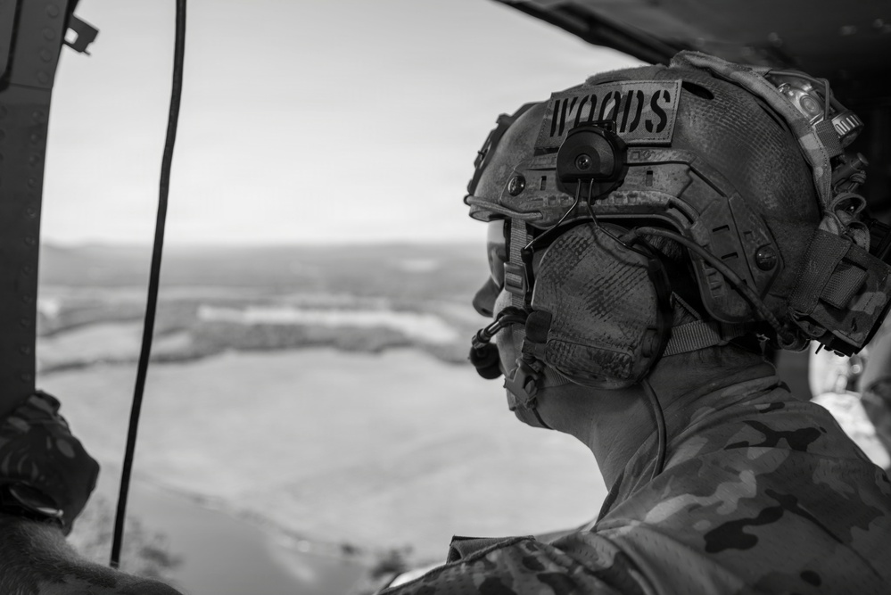 A black-and-white photograph of a military personnel identified by a name tape reading 'WOODS' on their helmet, viewed from inside what appears to be a helicopter. The individual is wearing full tactical gear including a combat helmet with mounted equipment, hearing protection, and camouflage uniform. They are looking out over a vast, flat terrain visible through the open aircraft door.